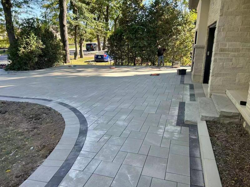 A curved interlock driveway in large-format grey pavers, edged with a charcoal banded border, being laid alongside a stone-clad home.