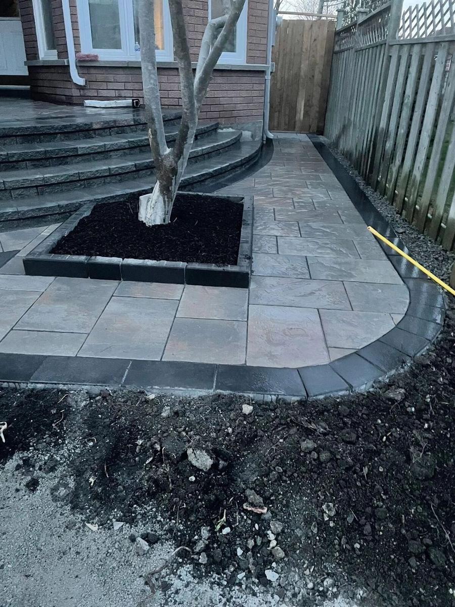 Curved paver patio with a tree well beside unfinished soil and wood steps
