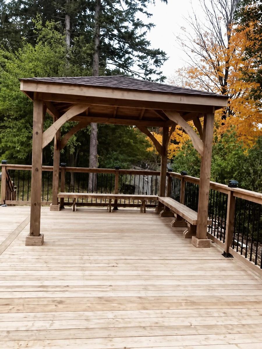 Timber gazebo with a shingled roof and built-in benches on a wood deck, backed by autumn trees