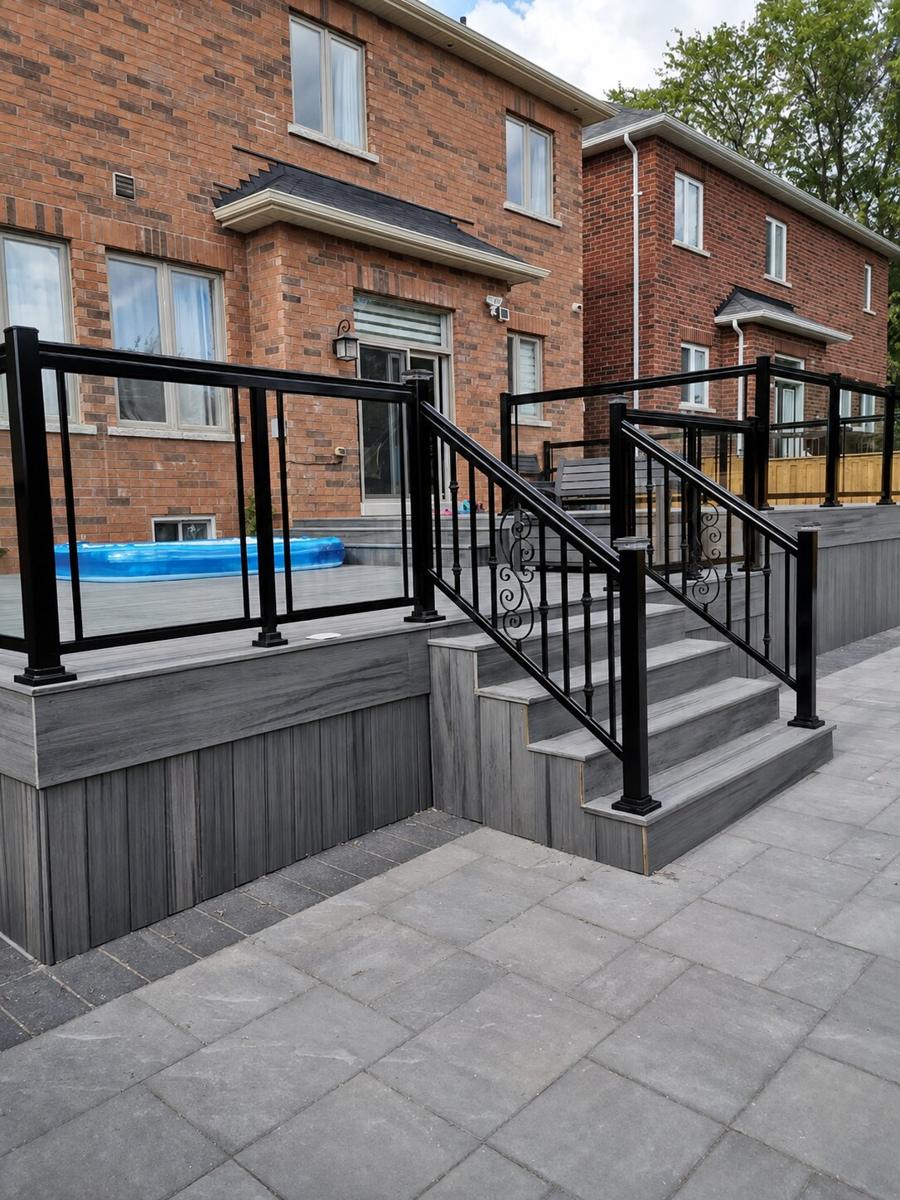 Grey deck with glass railings and decorative black metal stair balusters beside a brick home